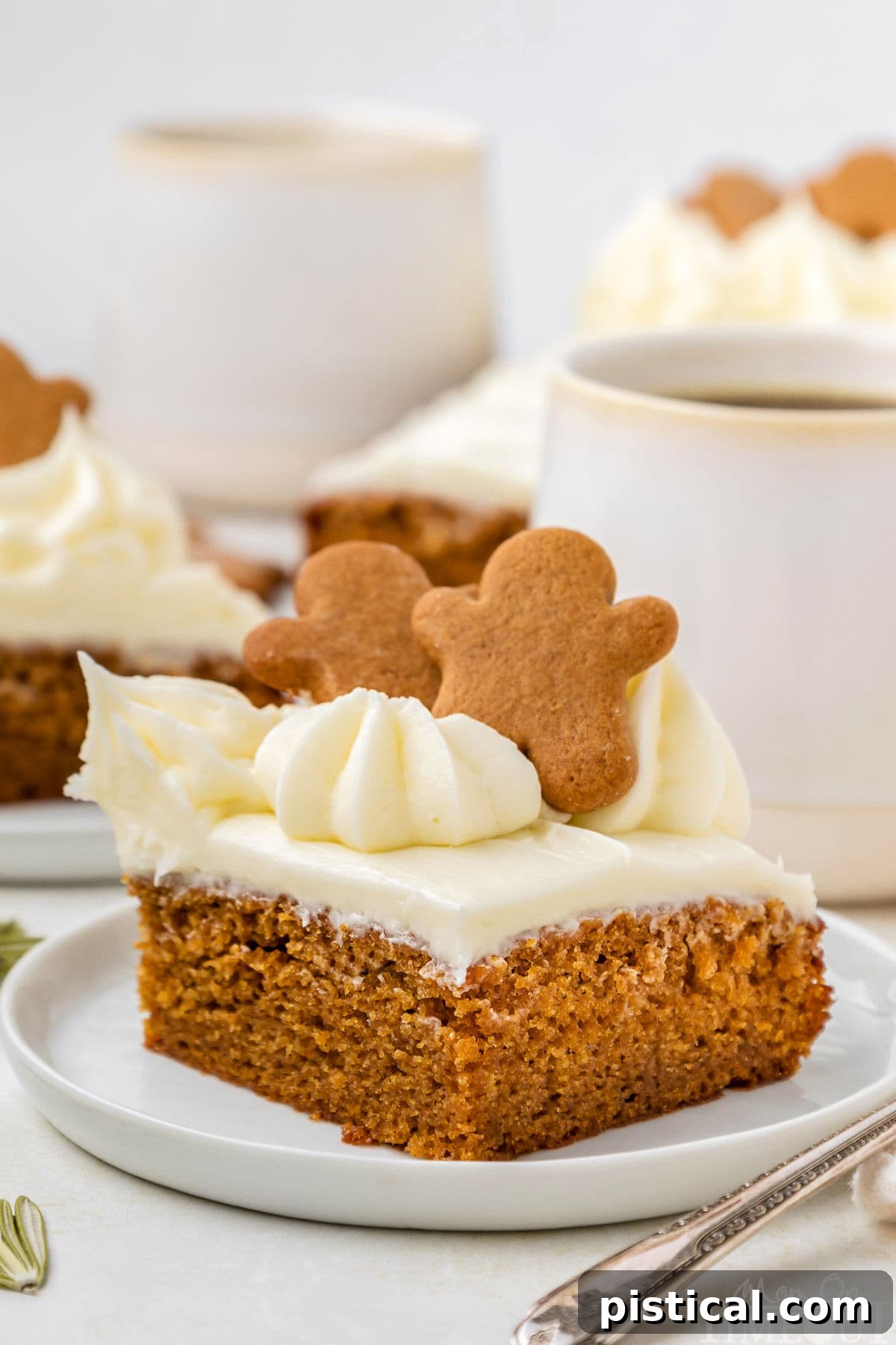 A slice of gingerbread cake topped with small gingerbread men. The cake sits on a white plate, with mugs of coffee and additional cake slices blurred in the background, creating a warm, inviting scene.