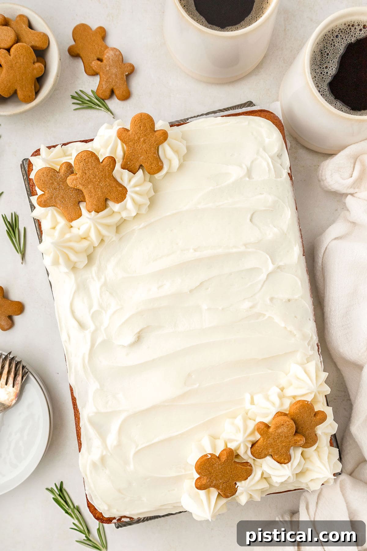 A top-down arrangement of a gingerbread cake on a white surface, surrounded by festive elements like coffee mugs, small gingerbread cookies, rosemary sprigs, plates, and a white linen. The composition is warm and inviting.