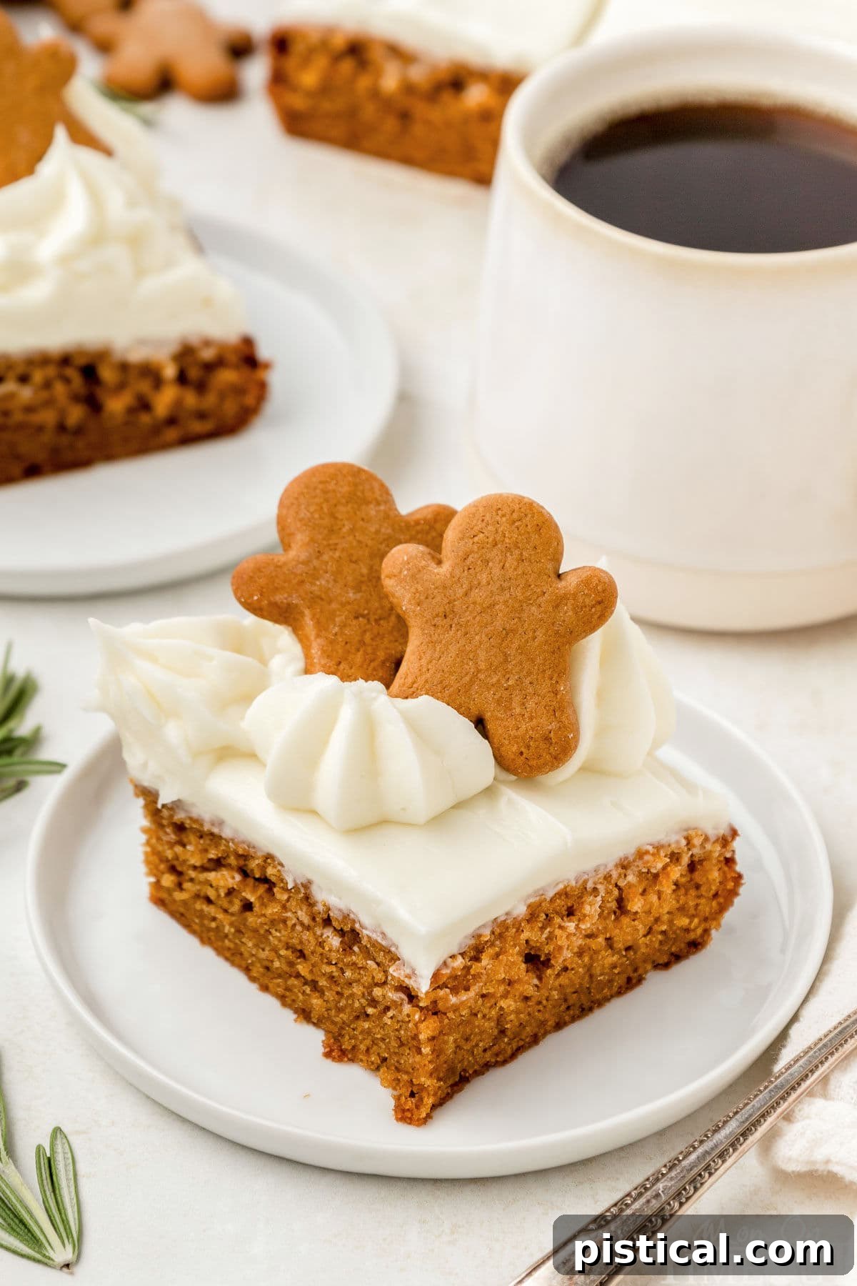 An angled shot looking down at a piece of gingerbread cake, elegantly frosted with cream cheese and placed on a round white plate. A warm cup of coffee is visible, blurred in the background, enhancing the cozy ambiance.