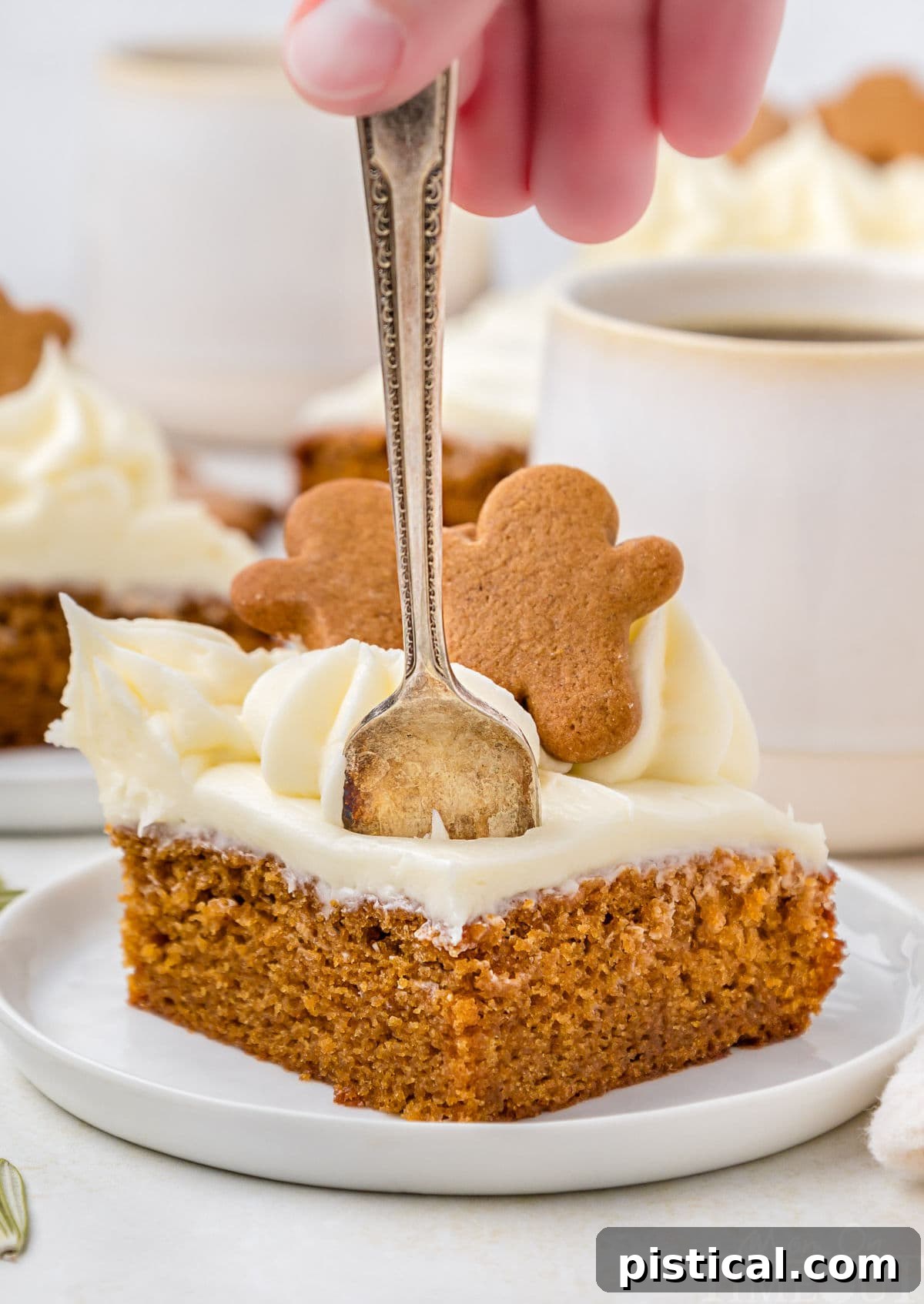 A hand gently inserts a fork into a tempting slice of gingerbread cake, which is perched on a white plate. In the softly blurred background, mugs of coffee and another slice of cake hint at a cozy, shared moment.