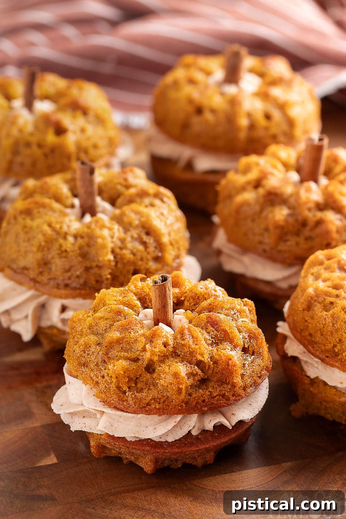 Six pumpkin bundt cakes with cinnamon stick stems sitting on dark wood board with striped towel in the background.
