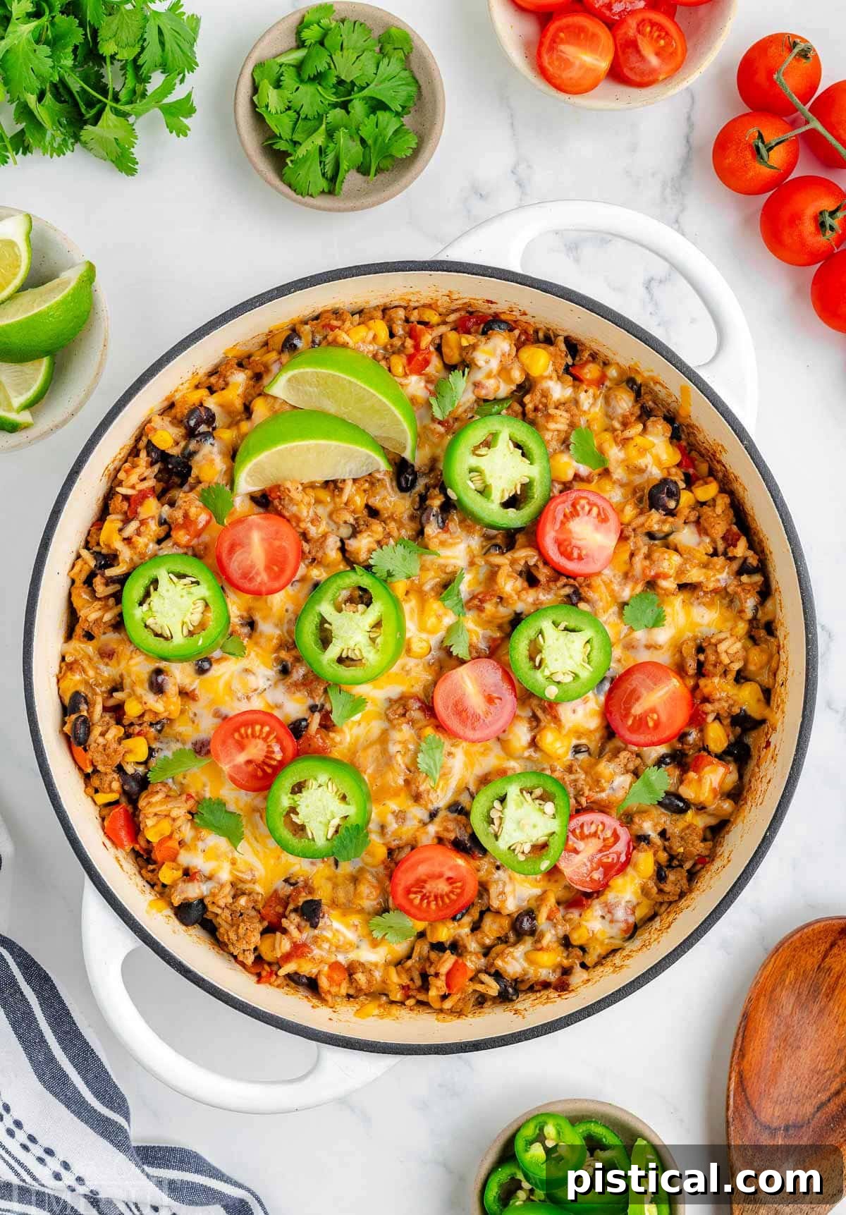 Ultimate Cheesy Taco Rice Skillet 2 A vibrant top-down view of Cheesy Taco Rice served in a white skillet, garnished with fresh jalapeño slices, bright lime wedges, and diced tomatoes. A scattering of various taco toppings like cilantro and avocado cubes are arranged enticingly around the skillet's edge, highlighting the dish's customizable appeal and rich textures.
