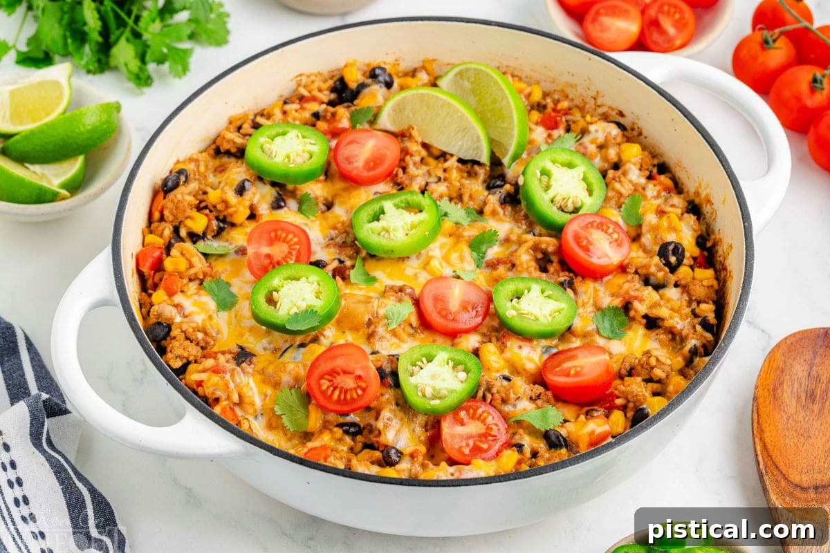 Ultimate Cheesy Taco Rice Skillet 6 An inviting top-down angled view of the Cheesy Taco Rice in a white skillet, adorned with fresh jalapeño slices, lime wedges, and diced tomatoes. The rich texture of the rice and melted cheese is visible, with a border of colorful taco toppings scattered around, enhancing its family-friendly appeal.