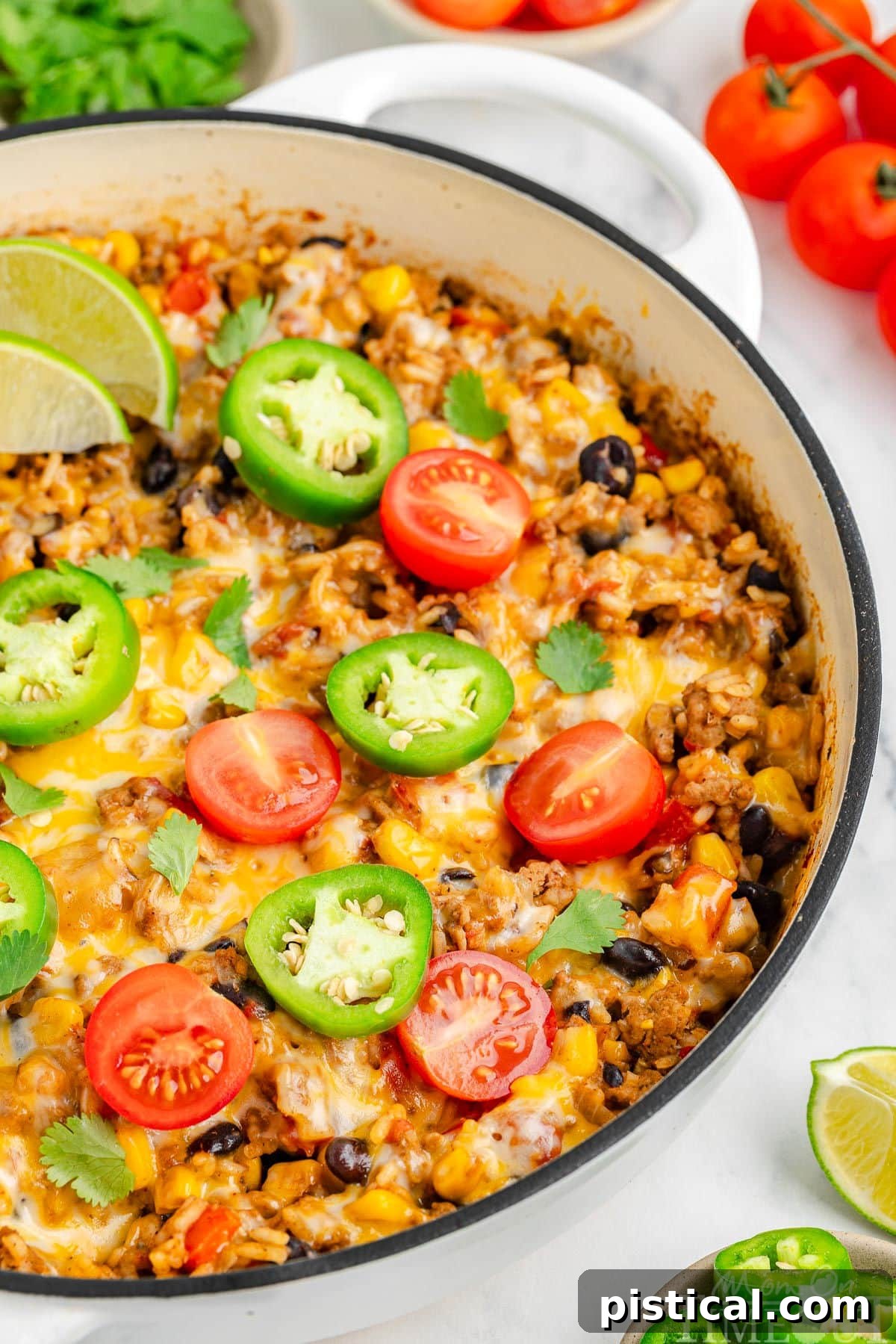 Ultimate Cheesy Taco Rice Skillet 7 A vibrant close-up view of Cheesy Taco Rice served directly in a white skillet, adorned with fresh jalapeño slices, lime wedges, and diced tomatoes. The rich, melty cheese and tender rice are prominently featured, with a scattering of various taco toppings around the edge, inviting a delicious spoonful.