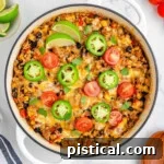 Ultimate Cheesy Taco Rice Skillet 8 Top down view of taco rice in a white skillet topped with jalapeno slices, lime wedges and tomatoes. More taco topping can be seen scattered around the edge of the skillet.