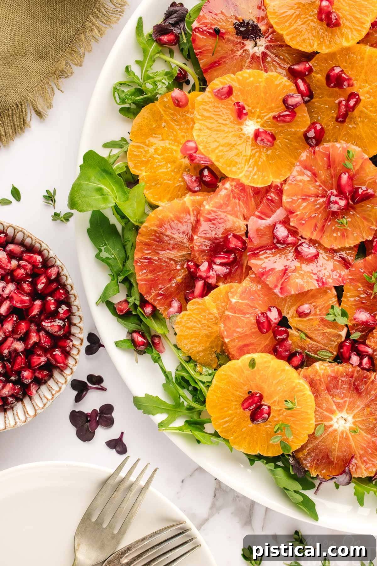 Winter citrus salad elegantly plated on large white platter and topped with thyme and pomegranate seeds. Oranges are cut in half and scattered around the white backdrop.