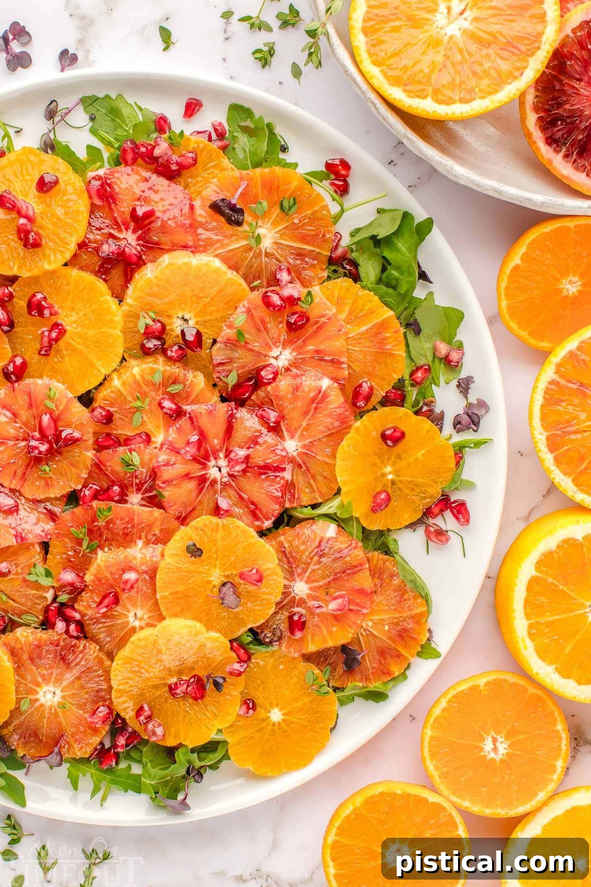 Winter citrus salad elegantly plated on large white platter and topped with thyme and pomegranate seeds. Oranges are cut in half and scattered around the white backdrop.
