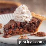Generous slice of chocolate bourbon pecan pie on white plate topped with whipped cream. The pie can be seen in the background.