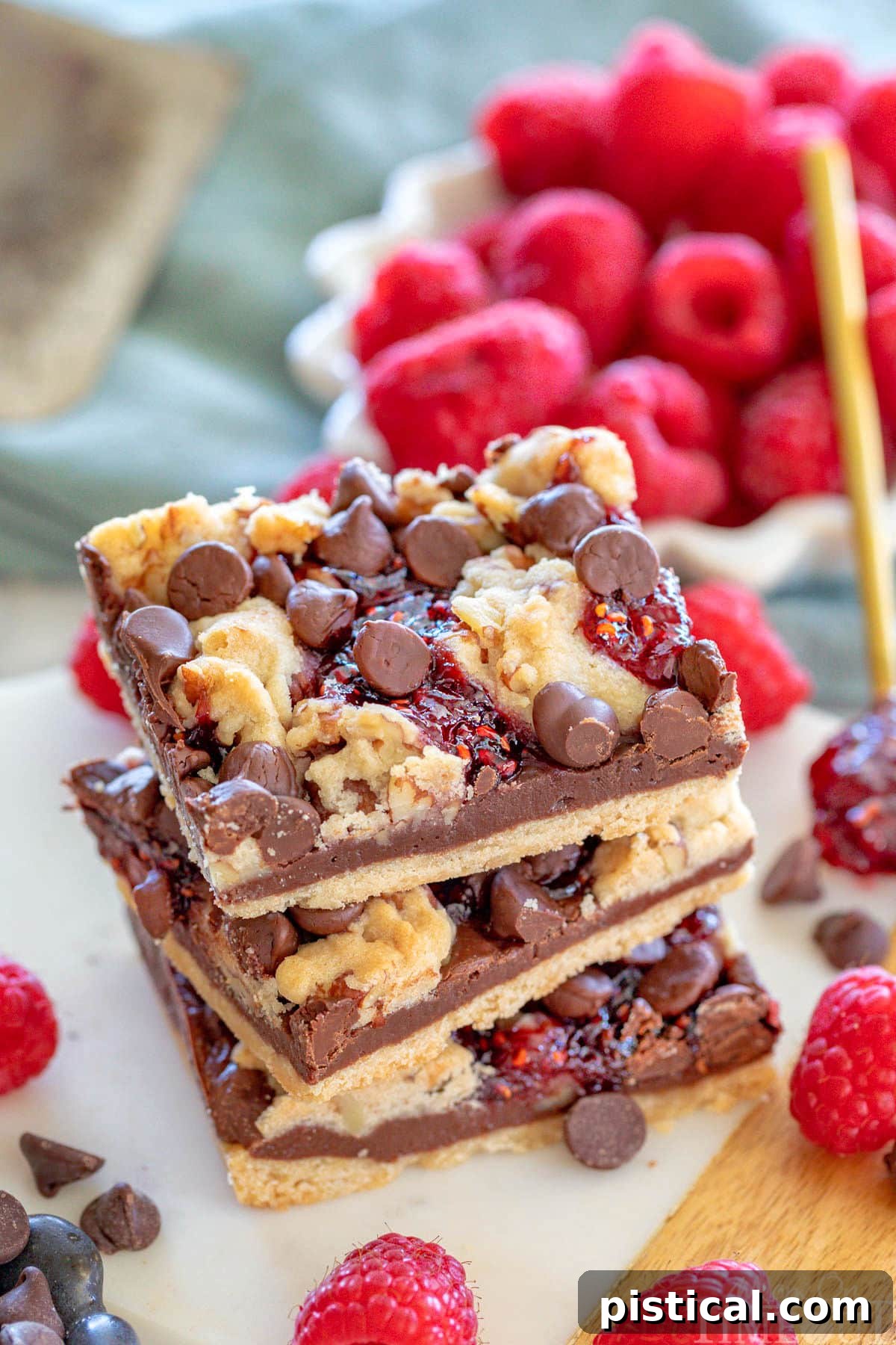 Three decadent chocolate raspberry crumb bars stacked on a white marble board, with fresh raspberries in a white bowl in the background.