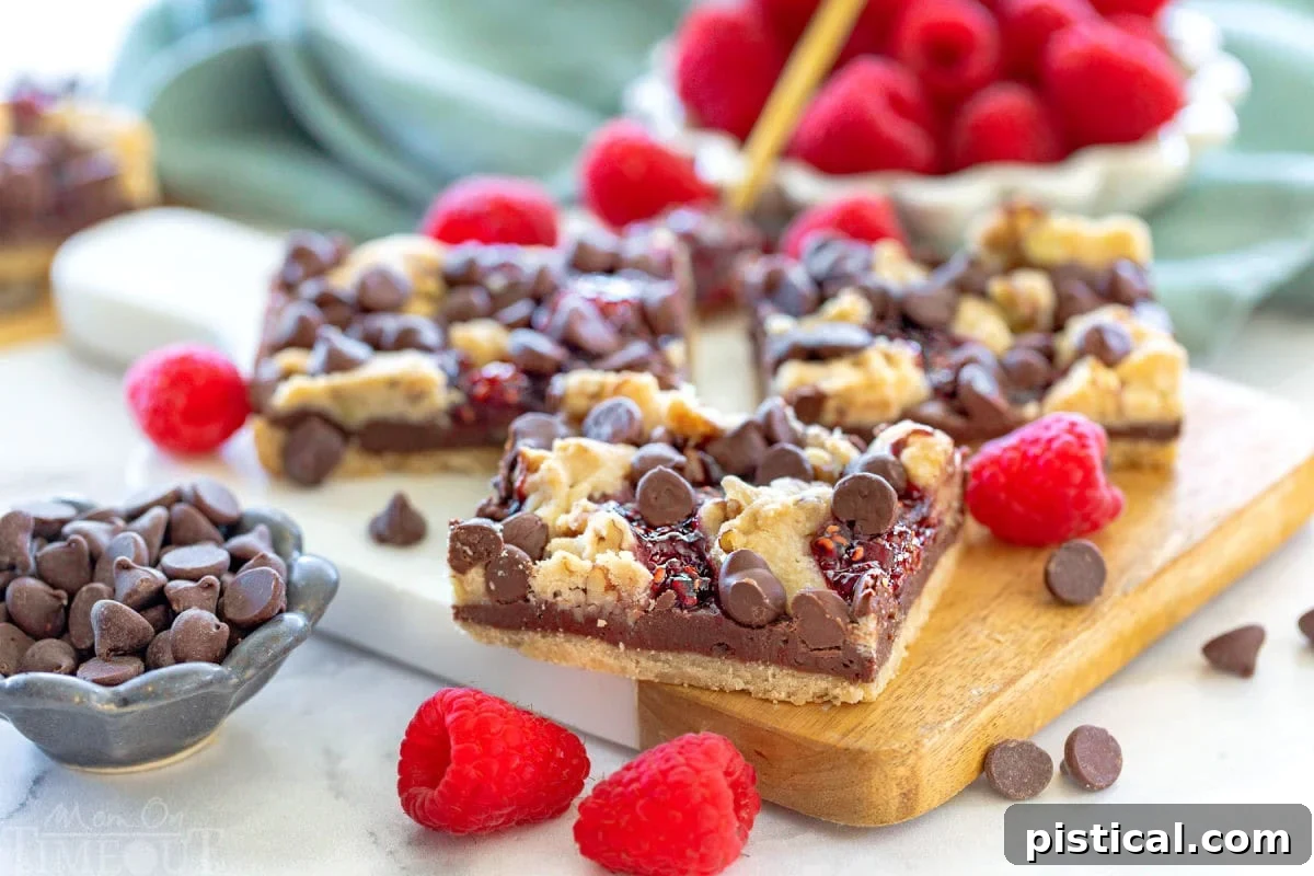 A wide shot of three chocolate raspberry crumb bars resting on a small white marble and wood cutting board, artfully arranged with chocolate chips and fresh raspberries sprinkled around them.