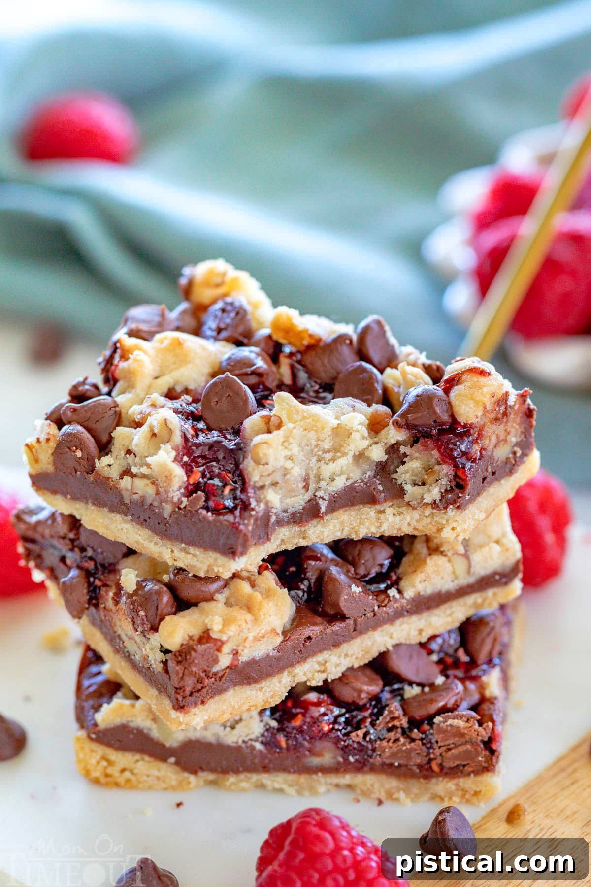 Three chocolate raspberry crumb bars stacked on a white marble board, with fresh raspberries in a white bowl in the background. The top bar has a bite delightfully taken from it, revealing the layers within.