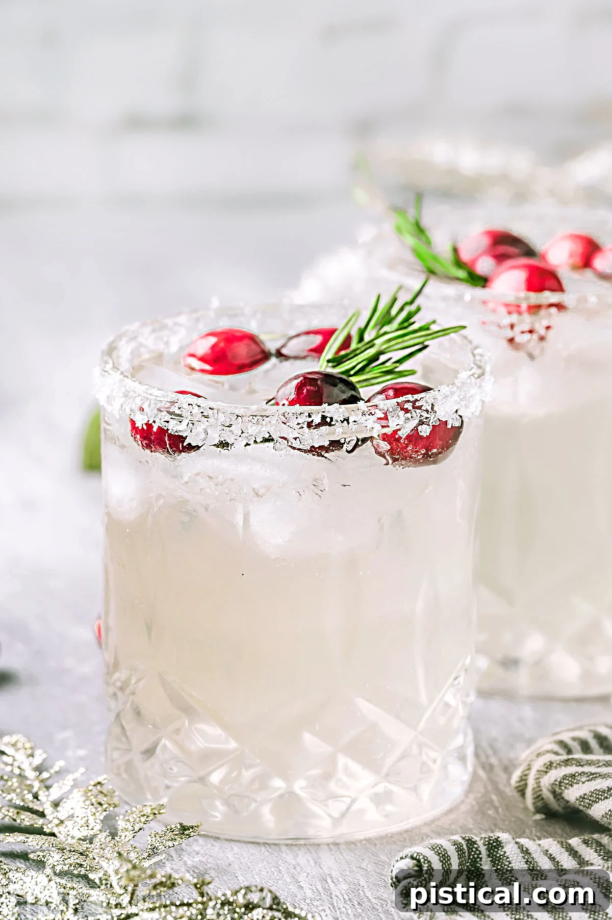 Two white christmas margaritas pictured on a white surface topped with cranberries and rosemary garnishes.