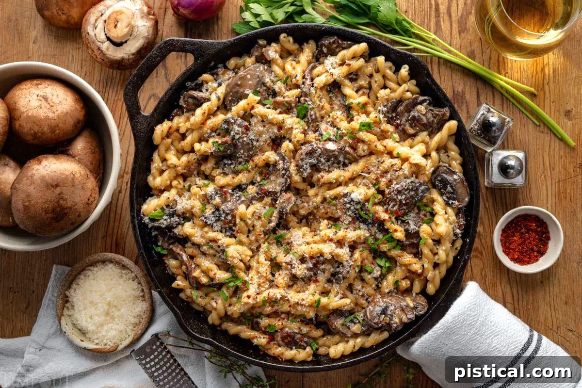 Top down view of cast iron skillet filled with mushroom pasta and finished with fresh parsley. Skillet is sitting on wood surface with mushrooms, shallots and herbs next to it.