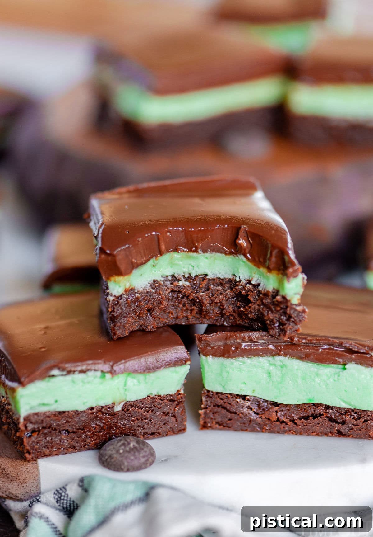 Three mint brownies stacked in a pyramid, with the top brownie having a bite taken out of it. More brownies are visible in the background, showcasing their irresistible layers.