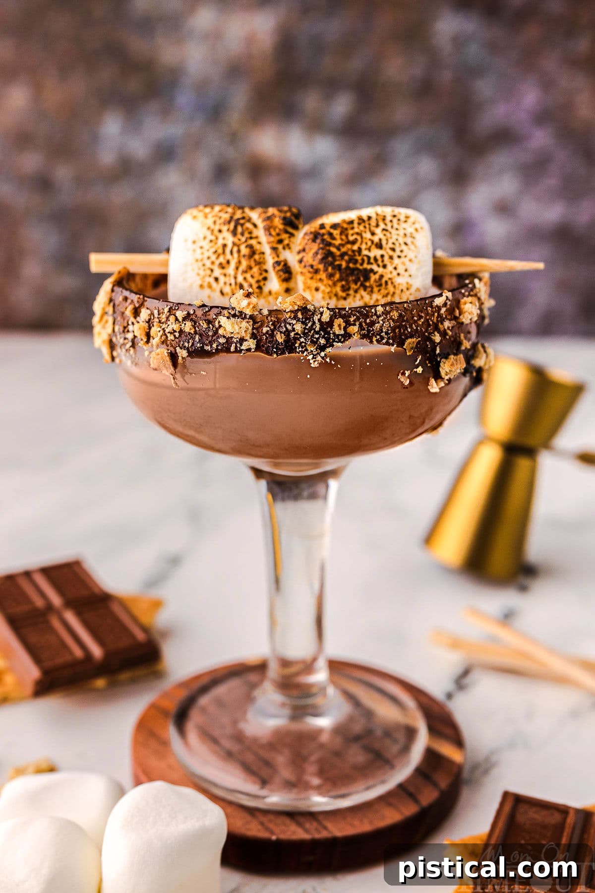 Sweet Campfire Martini 2 Side view of smores martini in a tall glass sitting on a wood coaster. Recipe ingredients are laying on the surface nearby.