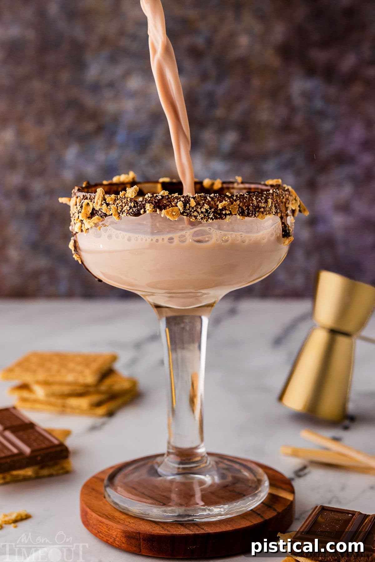Sweet Campfire Martini 3 Pouring smores martini into the glass that's on top of a wood coaster. Recipe ingredients are sitting on the surface nearby.