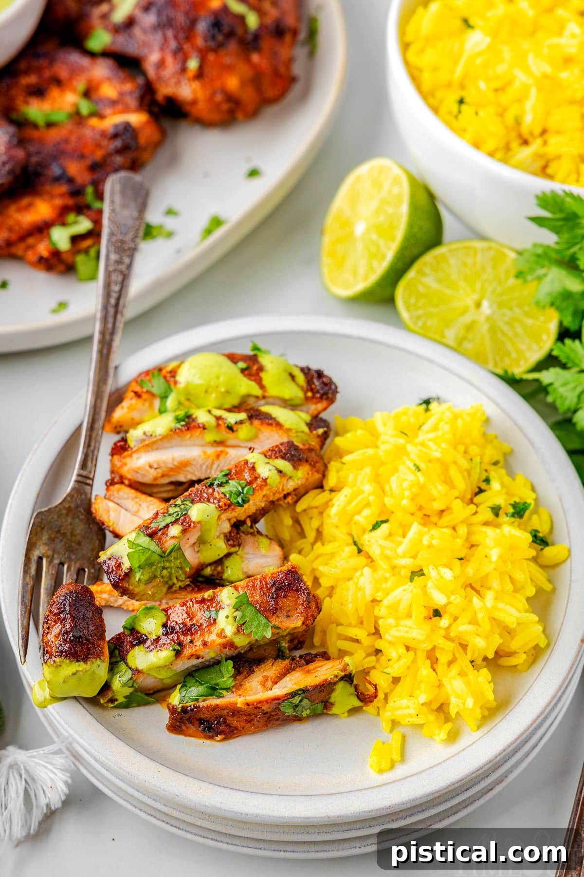 A top down view of sliced Peruvian Chicken next to a side of rice on a white plate with a fork holding a piece. Extra chicken and rice are on serving plates in the background.