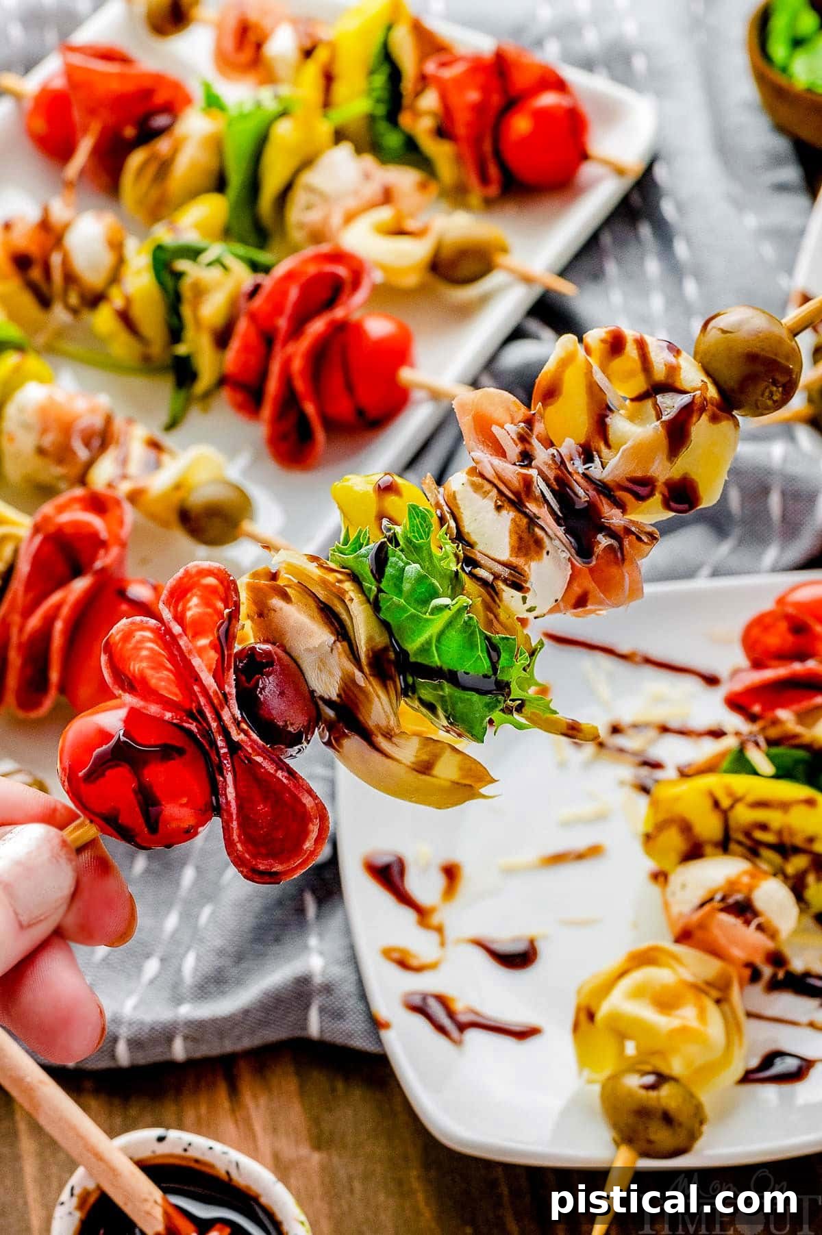 A hand holding up a single, perfectly assembled antipasto skewer, showcasing its colorful ingredients, over a blurred plate background.