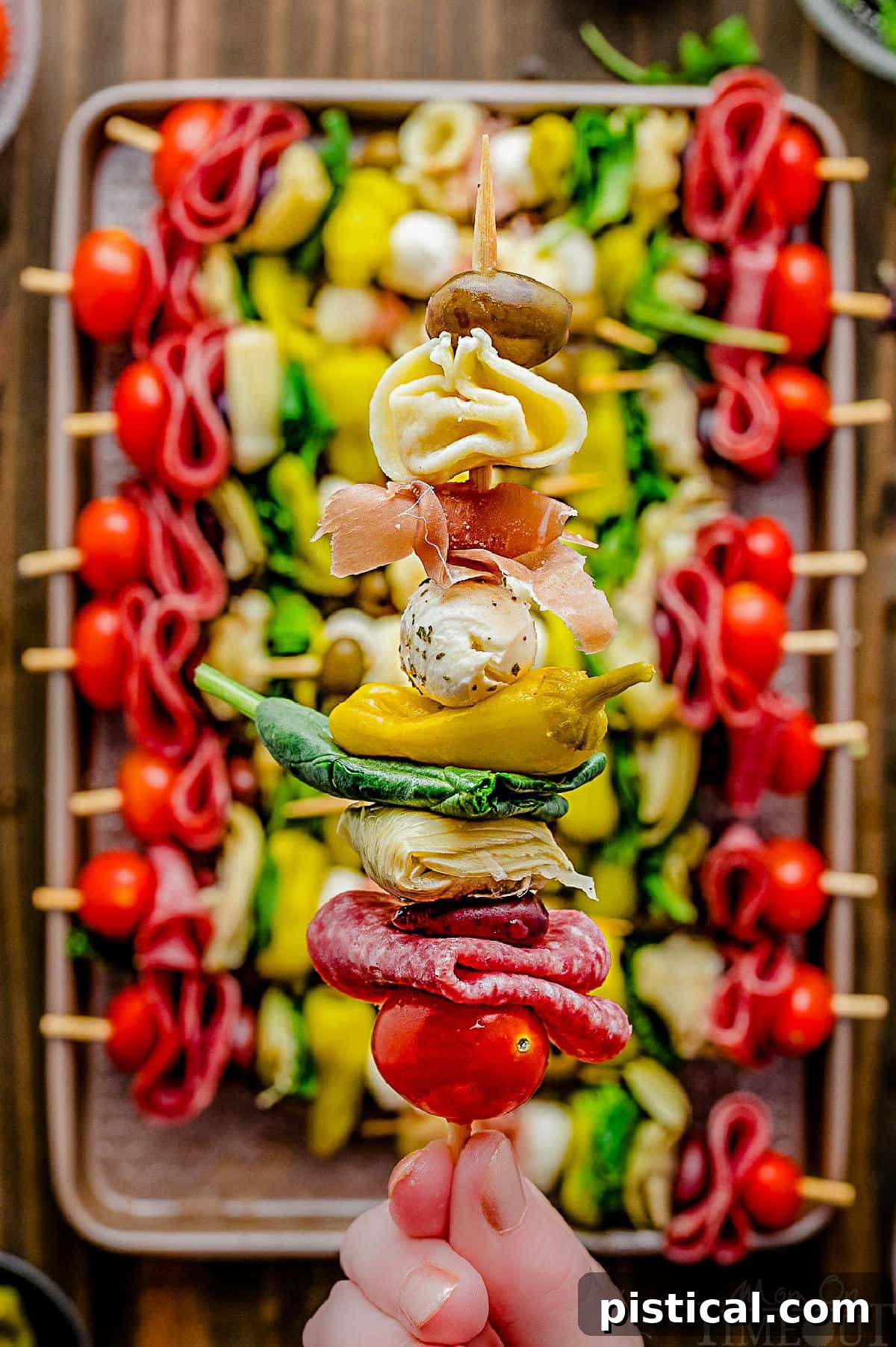 A single antipasto skewer held high above a sheet tray filled with many more colorful, freshly assembled skewers.
