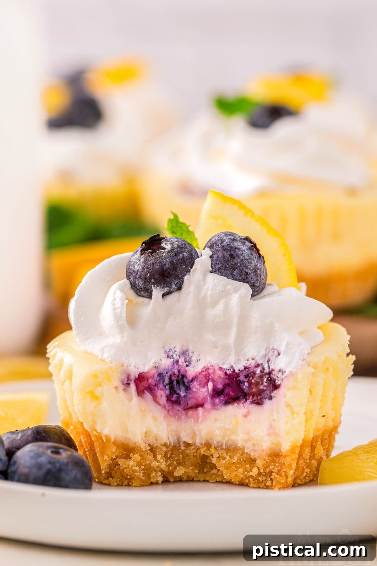 A close up view of a lemon blueberry mini cheesecake with a bite taken out of it. The cheesecake is on a white plate next to garnishes.