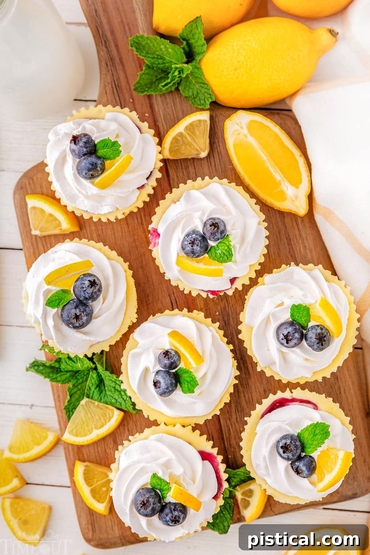 A top down view of lemon blueberry mini cheesecakes on a wood cutting board. The cutting board also has mint leaves and lemon slice garnishes on it.