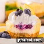A close up view of a lemon blueberry mini cheesecake with a bite taken out of it. The cheesecake is on a white plate next to garnishes.