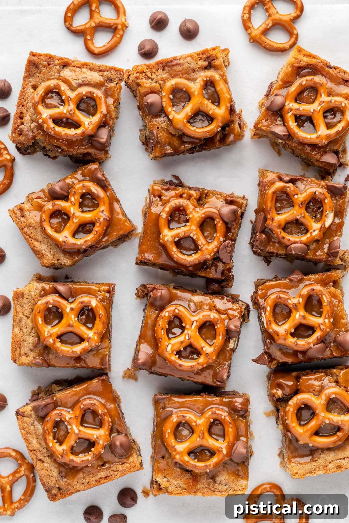 Sweet Salty Blondie Squares 3 Salted Caramel Pretzel Blondies lined up on a piece of parchment paper. Pretzels and chocolate chips are scattered around the bars.