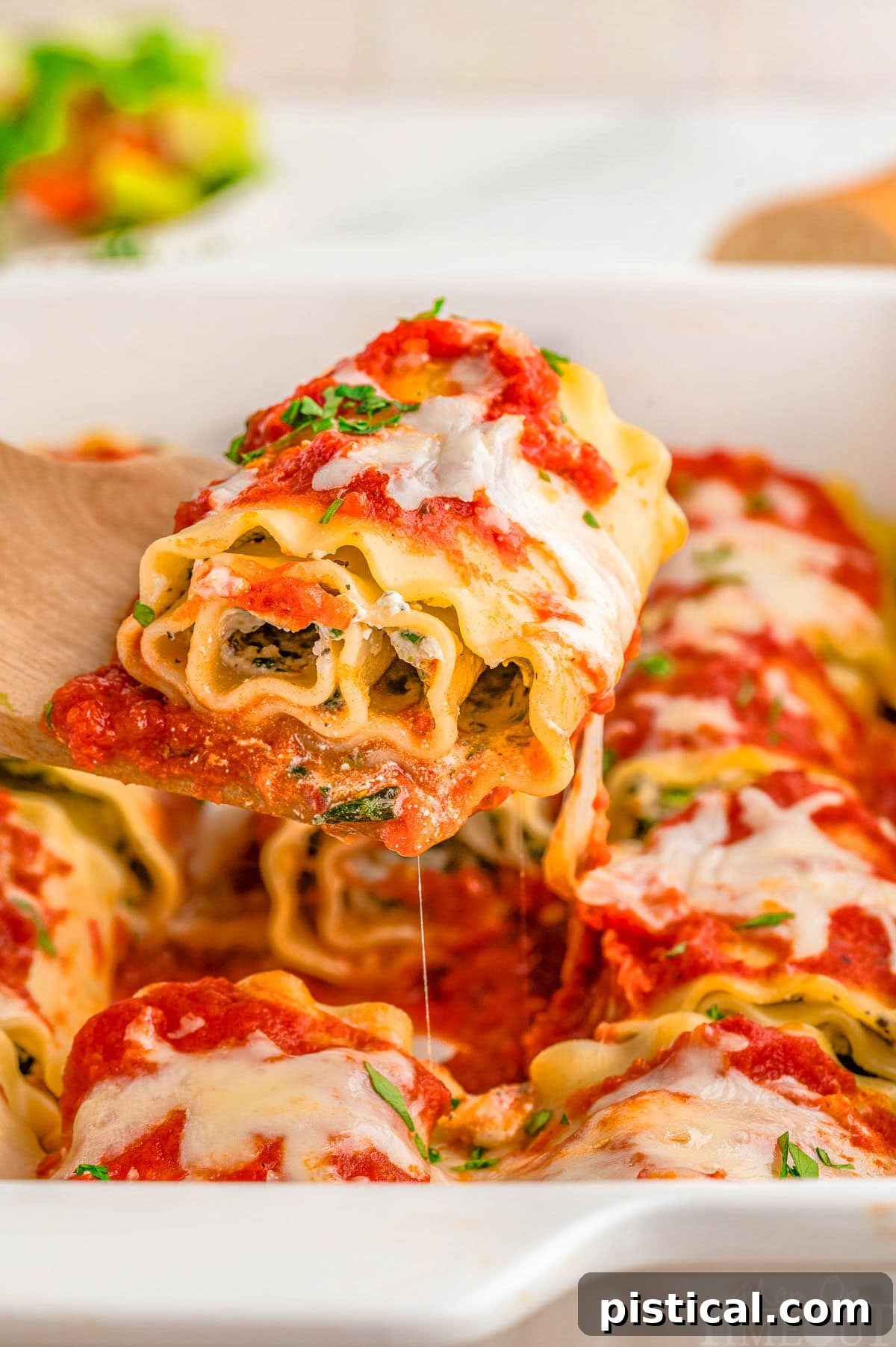 Lasagna roll up being lifted from baking dish with a wood spoon.