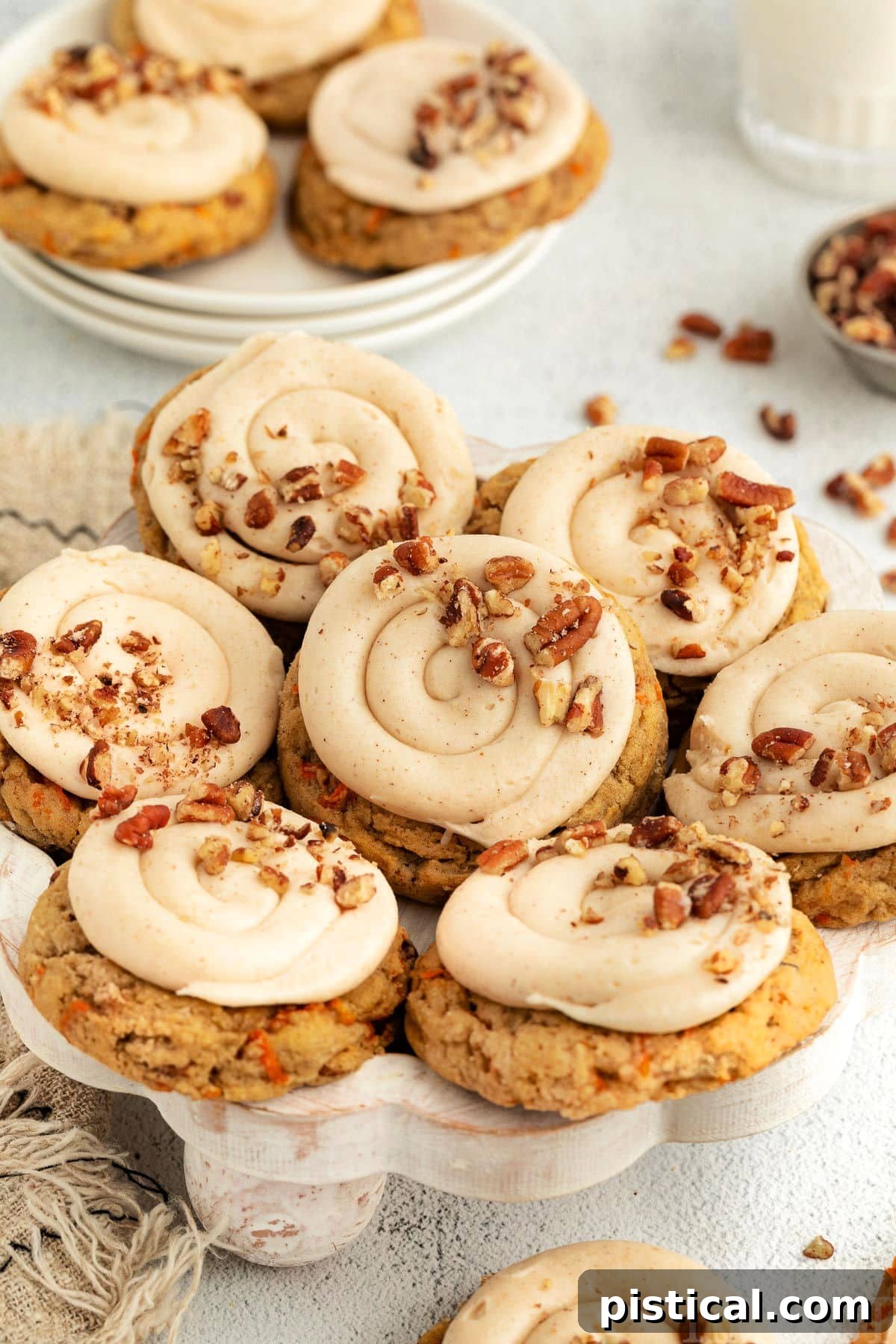 Cream Cheese Frosted Carrot Cookies 9 A collection of beautifully frosted Carrot Cake Cookies displayed on a pristine white wooden cake stand, with additional cookies and scattered pecans in the background, creating an inviting scene.