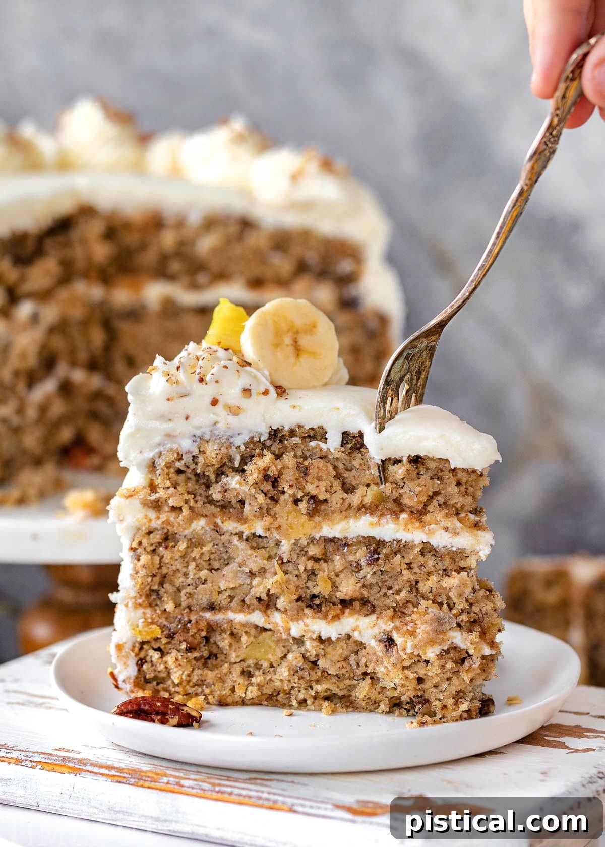 A perfectly sliced piece of Hummingbird Cake, showcasing its moist layers, rich cream cheese frosting, and visible flecks of banana and pineapple. A fork gently cuts into the top of the slice, with the rest of the beautifully frosted cake in the soft background.