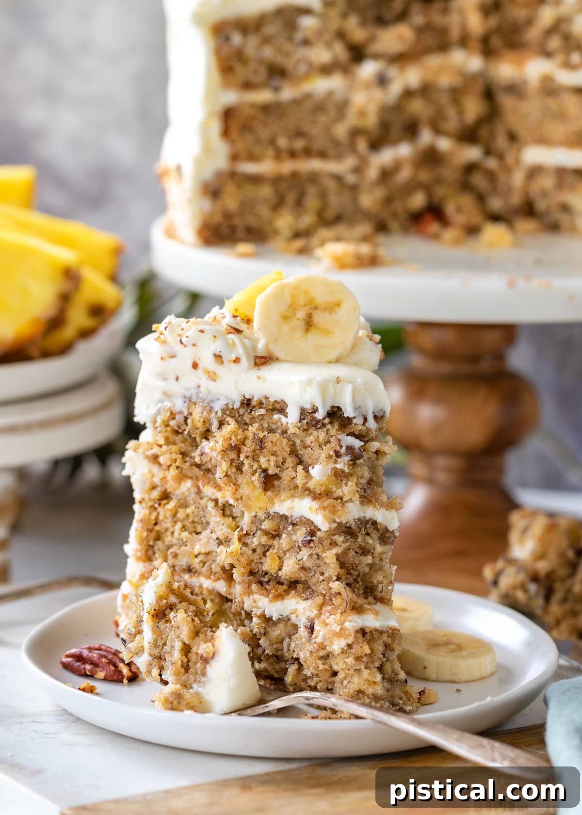 A single, inviting slice of Hummingbird Cake, beautifully presented on a round white plate, with the majestic full cake resting on a cake stand in the background, ready to be enjoyed.