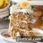 Slice of hummingbird cake recipe on a white round plate sitting in front of cake stand with rest of cake on it.