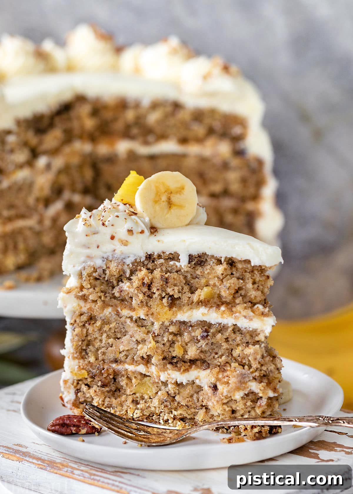 A delectable slice of Hummingbird Cake, artfully topped with banana and pineapple garnishes, and generously frosted with cream cheese. The larger, beautifully decorated cake is visible in the background.