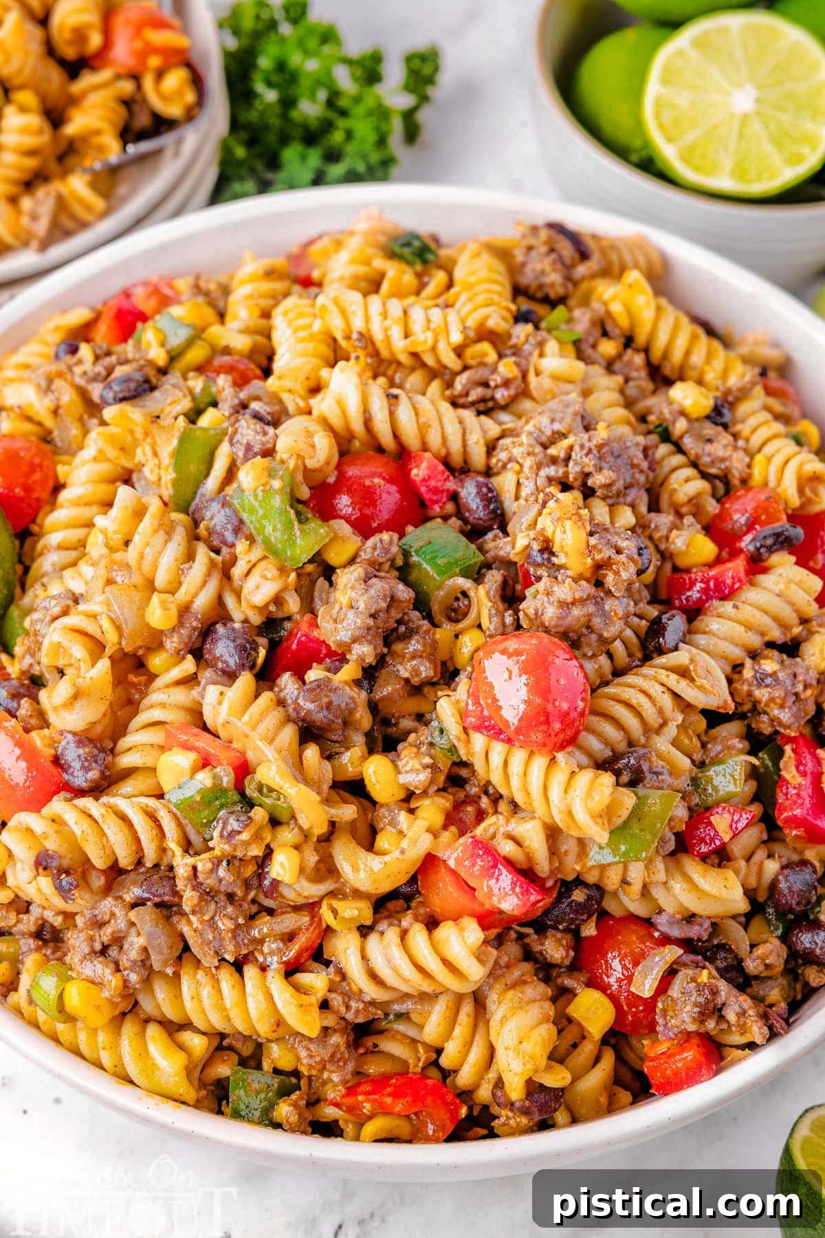 Ranch Hand Pasta Salad 2 A large serving bowl overflowing with vibrant Cowboy Pasta Salad, featuring pasta, ground beef, colorful vegetables, and a zesty dressing, ready to be served. In the background, a plate of the salad is visible alongside fresh lime wedges.