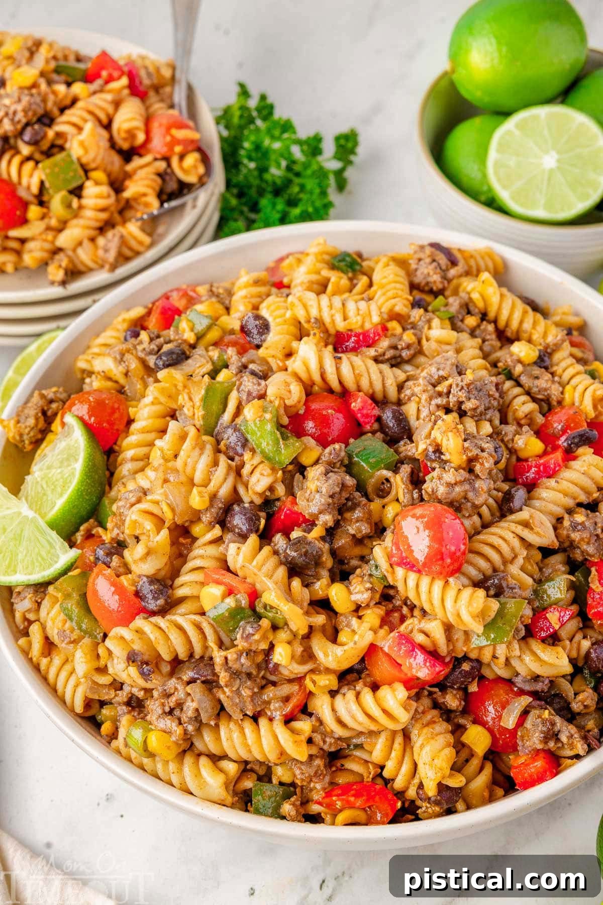 Ranch Hand Pasta Salad 8 A beautifully presented Cowboy Pasta Salad in a pristine white serving bowl, elegantly garnished with fresh lime wedges. In the softly blurred background, a stack of clean plates topped with portions of the same inviting salad suggests a meal ready for enjoyment.
