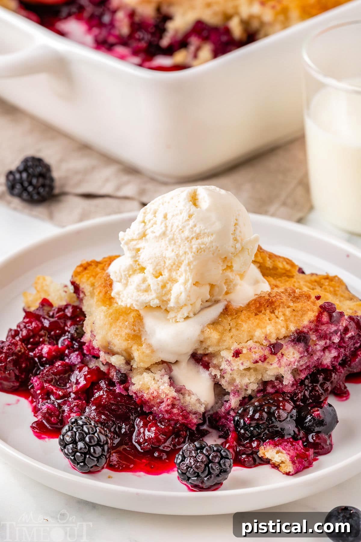 Rustic Berry Bake 2 A serving of homemade Berry Cobbler topped with melting vanilla ice cream on a white plate. In the background, the baking dish holds the remaining cobbler.