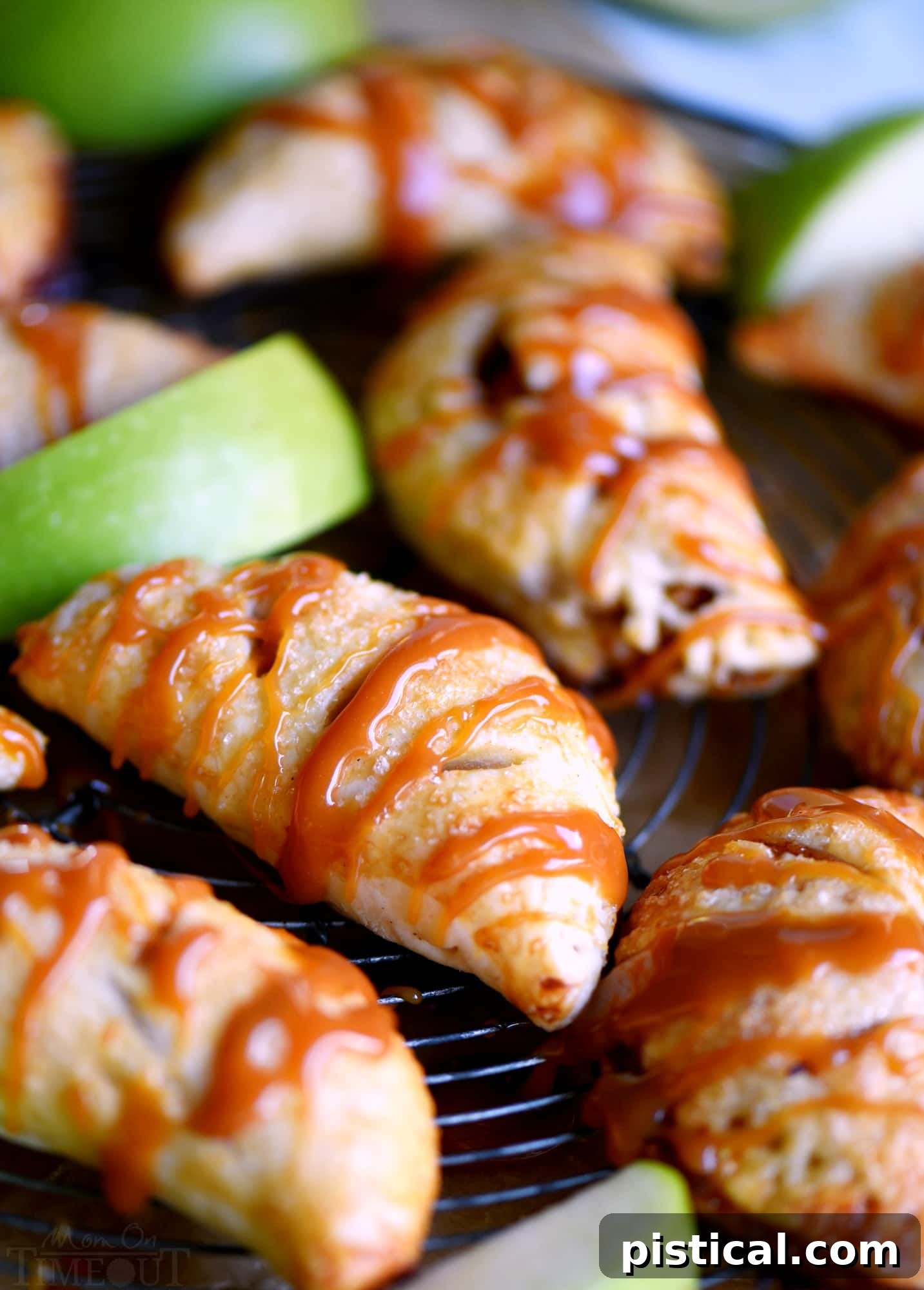 A stack of golden brown caramel apple empanadas, some cut in half to reveal the apple and caramel filling, all beautifully drizzled with extra caramel sauce.