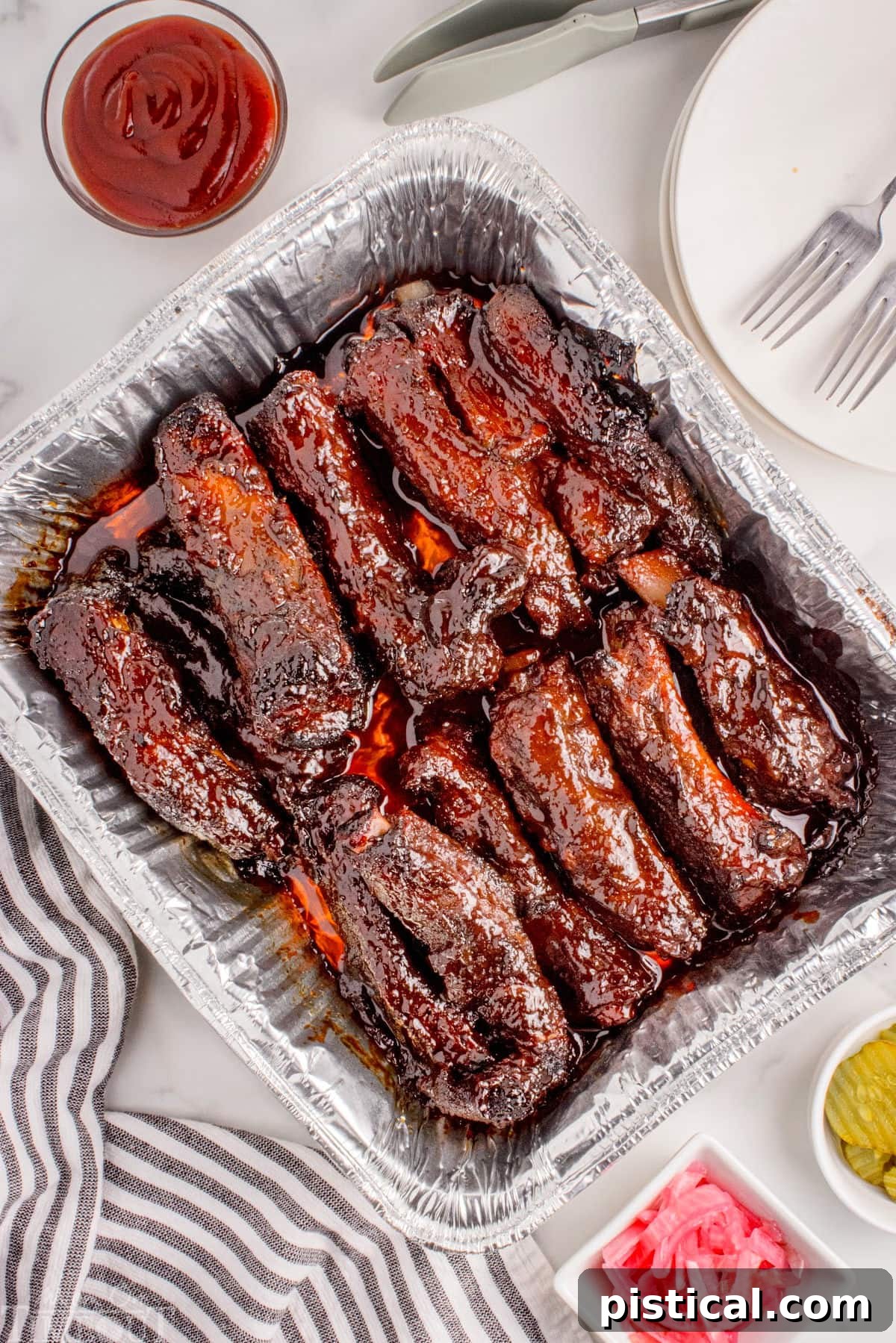 Overhead shot of grilled party ribs in foil pan with bbq sauce, pickled onions and pickles in small bowls to the side.