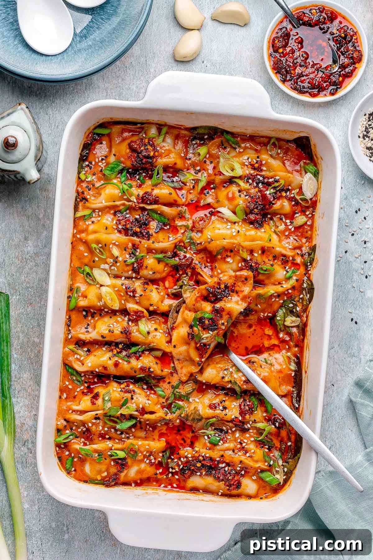 Crispy Sheet Pan Dumplings 2 Overhead shot of vibrant baked dumplings in a white baking dish, garnished with chili crisp, fresh green onions, and a sprinkle of black and white sesame seeds. A spoon is poised in the dish, ready to serve this delightful meal.