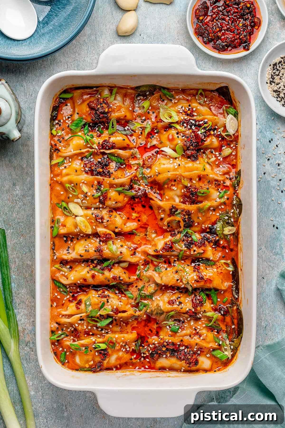 Crispy Sheet Pan Dumplings 9 Overhead shot of a white baking dish filled with viral baked dumplings, beautifully topped with fiery chili crisp, fresh green onions, and a sprinkle of contrasting black and white sesame seeds. A spoon is strategically placed, ready for serving.