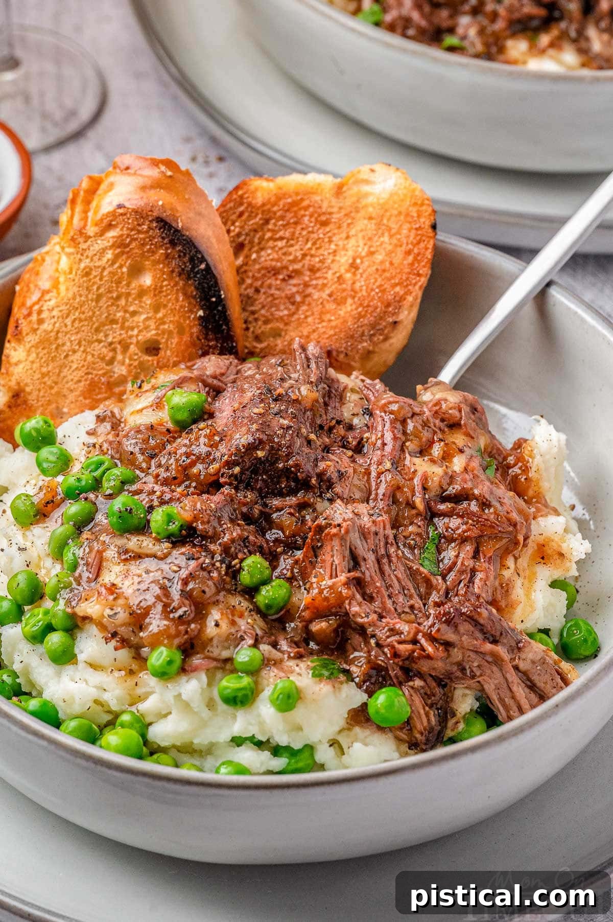 Close up shot of French Onion Soup Pot Roast served in a bowl with creamy mashed potatoes, green peas, and a piece of toasted French bread, highlighting the rich texture.