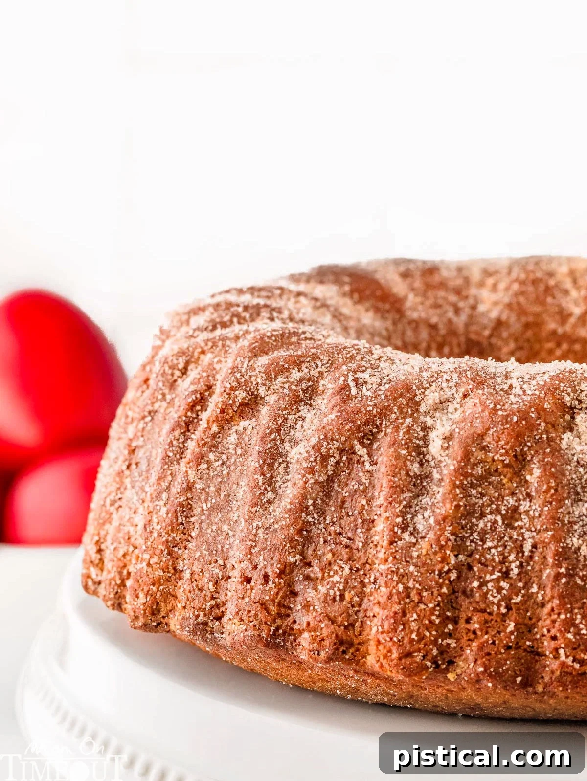 A stunning side view of a whole Apple Cider Bundt Cake, glistening with its cinnamon-sugar coating, elegantly displayed on a white cake stand with vibrant red apples artfully arranged in the background, signaling the heart of autumn.