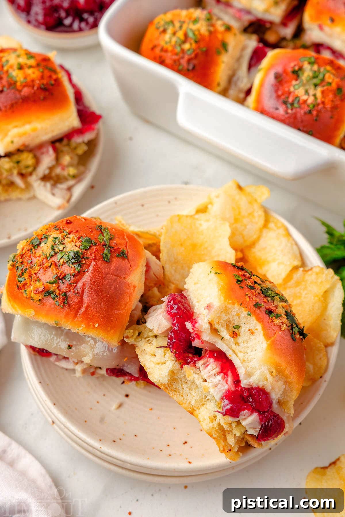 Two turkey sliders, one with a bite taken out, served on a white plate with potato chips. The baking dish with remaining sliders is blurred in the background.