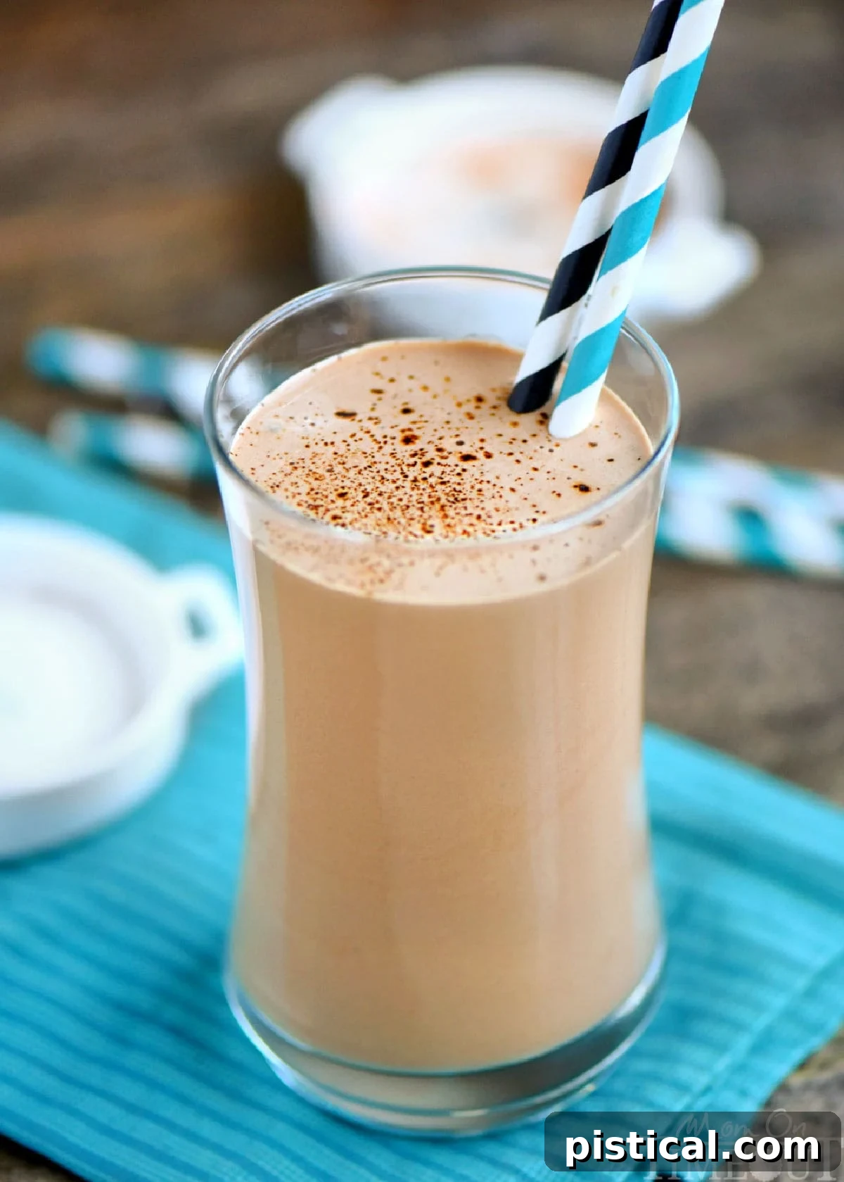 A tall glass filled with a creamy coffee protein shake, garnished with two eco-friendly paper straws, resting on a stylish teal napkin atop a warm wooden surface. The perfect energizing start to your day.