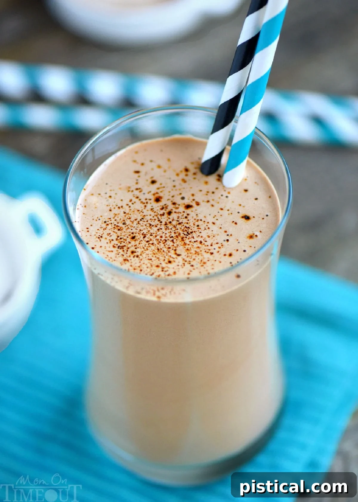 An inviting angled top-down view of a creamy protein and coffee shake, elegantly poured into a tall glass and finished with a delicate dusting of espresso powder. The glass rests on a striped teal towel, with a few extra straws in the background, suggesting a moment of delightful indulgence.