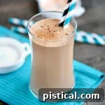 Coffee protein shake in a tall glass with two paper straws. Glass is sitting on teal napkin on a wood surface.