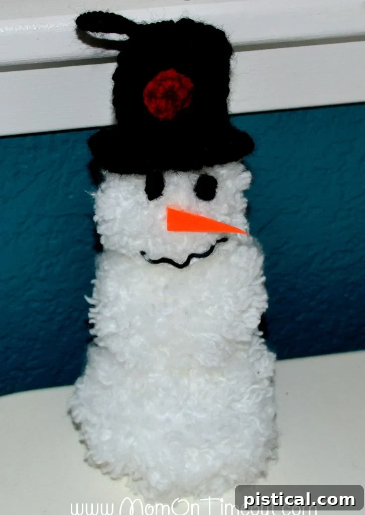 Two finished pom pom snowman ornaments, one with a red scarf and one with a patterned ribbon, hanging on a Christmas tree