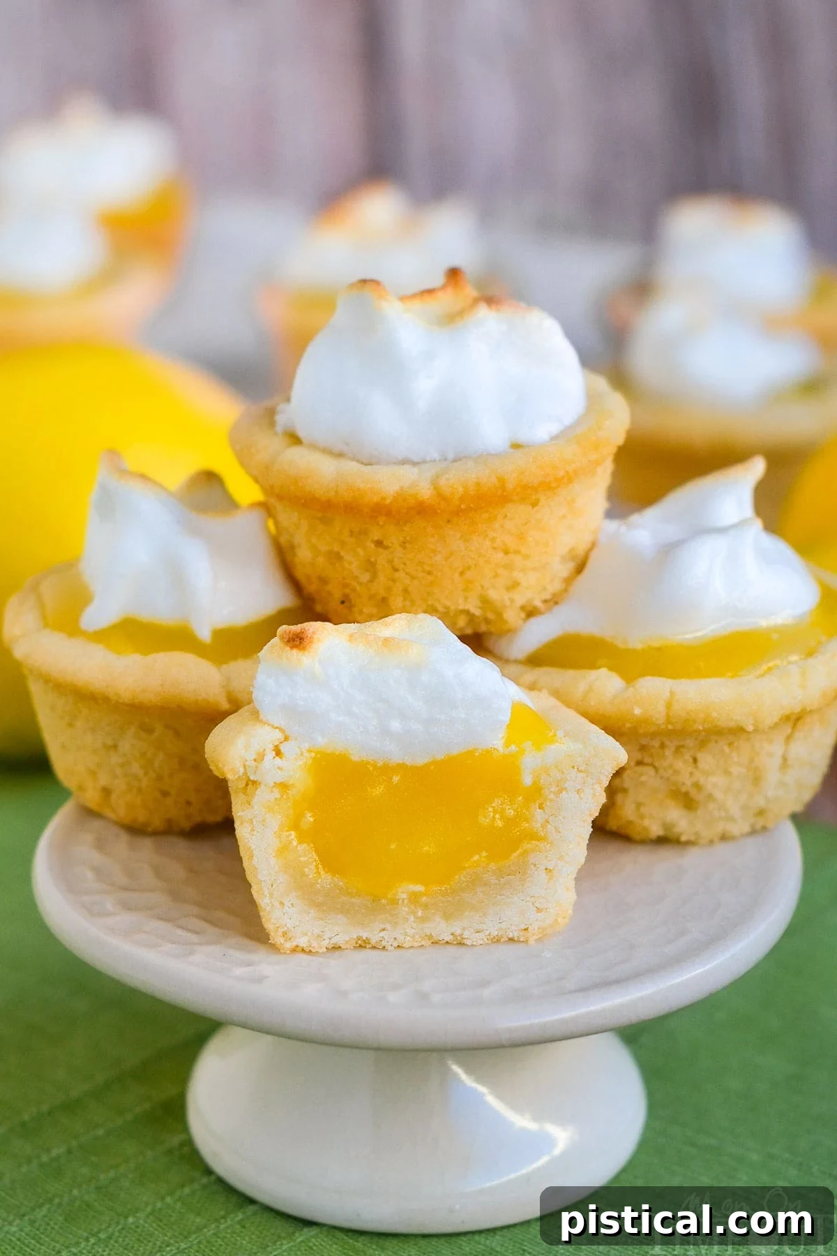 A stack of golden brown Lemon Meringue Cookie Cups, each topped with a delicate swirl of toasted meringue, arranged on a pristine white cake stand. The vibrant yellow lemon curd peeks out from beneath the meringue, hinting at the delightful tartness within. Perfect for a sunny gathering or a sweet escape.