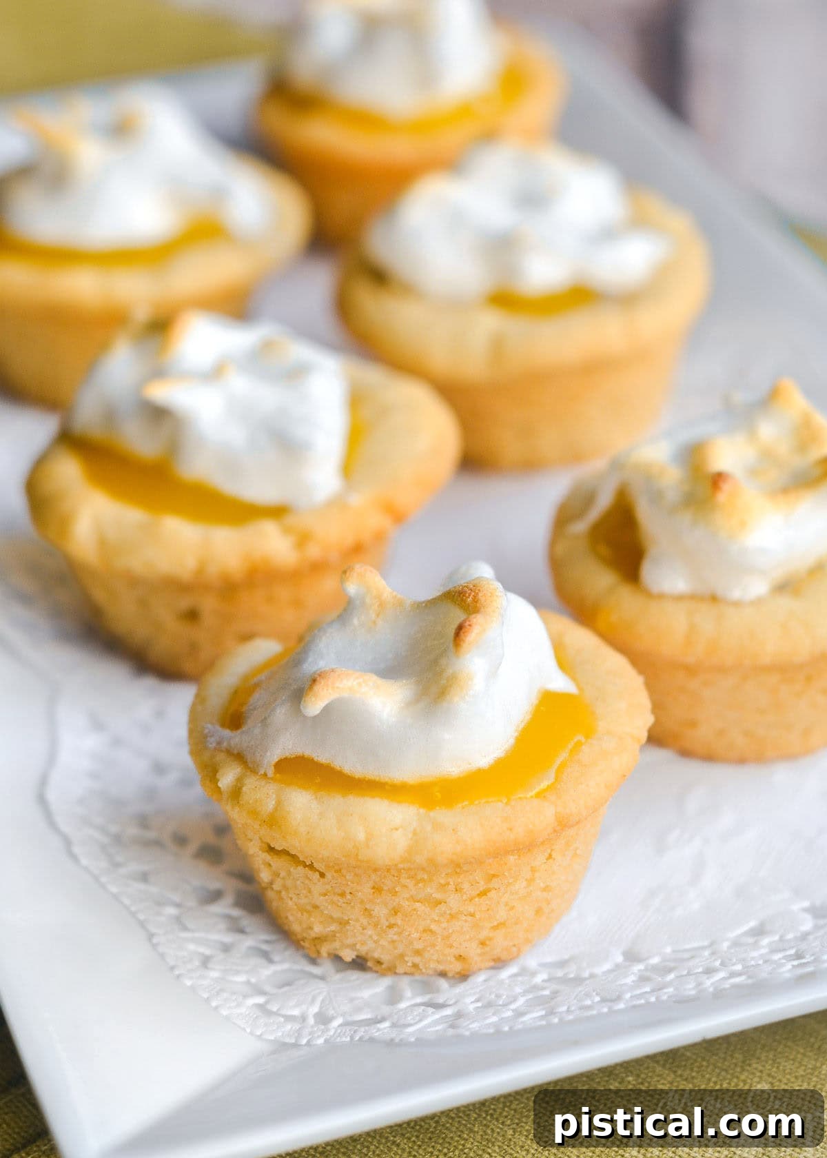 Individual lemon meringue cookie cups on a pristine white ceramic serving dish, highlighting their delicate toasted meringue peaks and golden cookie bases. The lighting is soft and inviting.