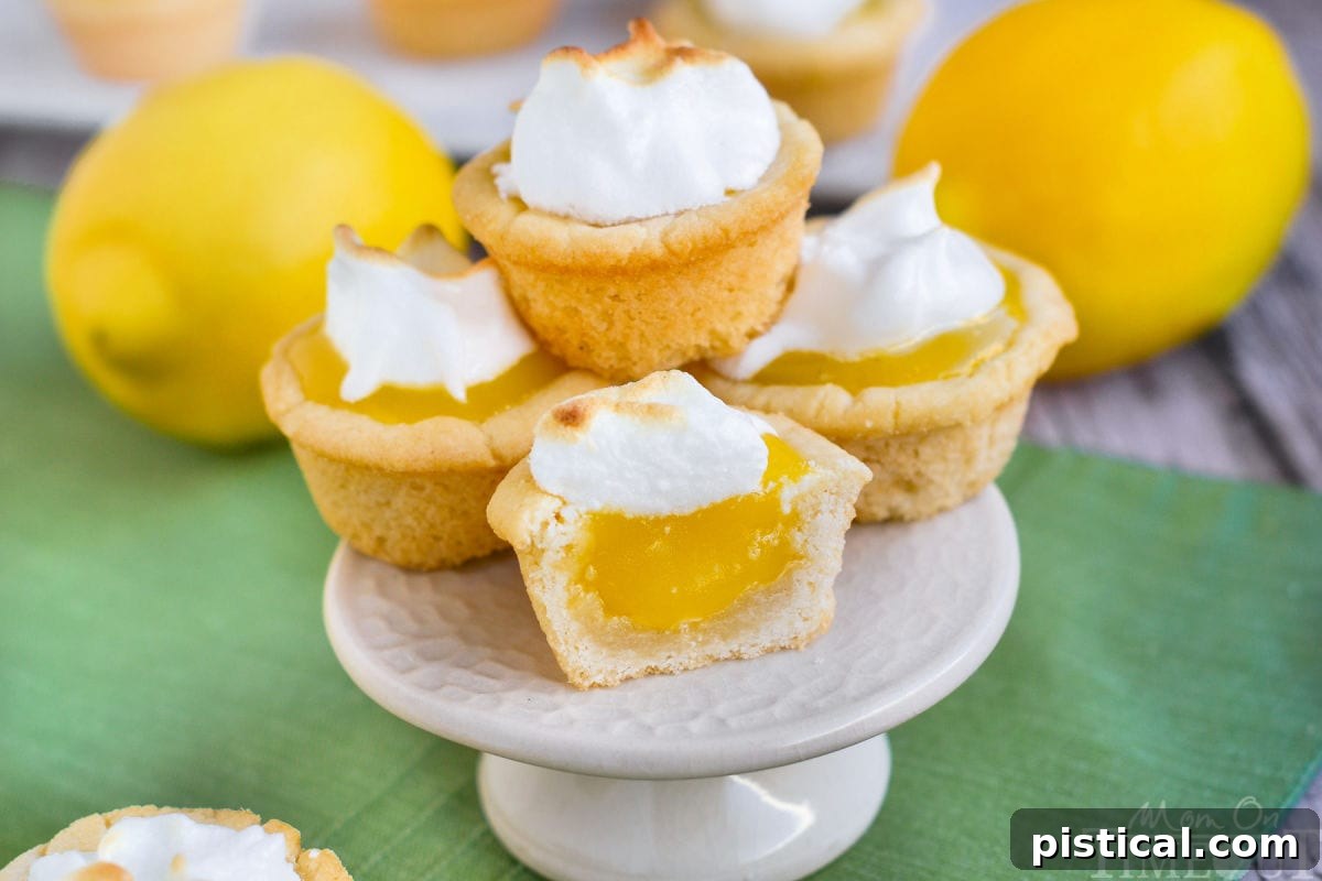 A stack of golden Lemon Meringue Cookie Cups, each adorned with a perfectly toasted meringue swirl, sits on a white cake stand. The vibrant lemon curd layer is visible beneath the fluffy topping.